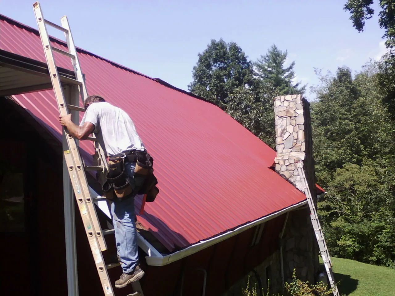 Roofer on a ladder beside a red metal roof and stone chimney — Soffit & Fascia Repair by Roofing Brothers in Crowley
